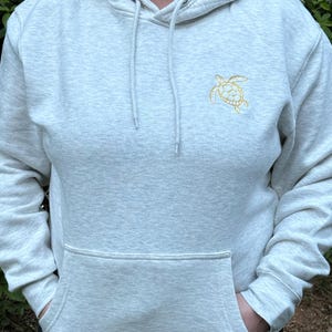 Puede incluir: Sudadera con capucha gris claro con bolsillo de canguro y capucha con cord&oacute;n. La sudadera tiene una peque&ntilde;a tortuga marina dorada bordada en la parte superior izquierda del pecho. La persona lleva pantalones negros.