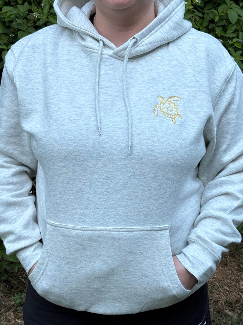 Puede incluir: Sudadera con capucha gris claro con bolsillo de canguro y capucha con cord&oacute;n. La sudadera tiene una peque&ntilde;a tortuga marina dorada bordada en la parte superior izquierda del pecho. La persona lleva pantalones negros.