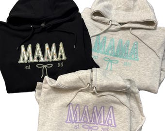 May include: Three hooded sweatshirts in black, light gray, and gray. Each sweatshirt has the word "MAMA" printed on the front, along with "est. 2025" and a bow graphic. The "MAMA" text is in different colors: green, teal, and purple.