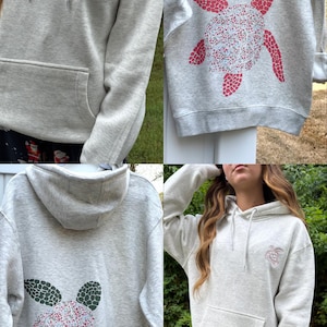 Puede incluir: Sudadera con capucha gris claro con bolsillo delantero y capucha. La sudadera presenta una peque&ntilde;a tortuga bordada en la parte delantera y un dise&ntilde;o de tortuga m&aacute;s grande en la parte trasera. Los dise&ntilde;os de tortugas tienen diferentes patrones de color.