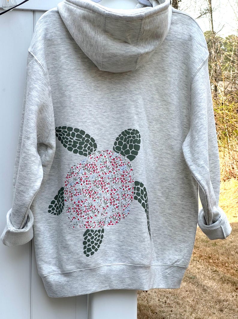 Puede incluir: Sudadera con capucha gris claro con un dise&ntilde;o de tortuga en la espalda. El caparaz&oacute;n de la tortuga est&aacute; relleno con un estampado floral de flores rojas, blancas y azules. Las aletas y la cabeza de la tortuga son de color verde oscuro. La sudadera tiene capucha y mangas largas.