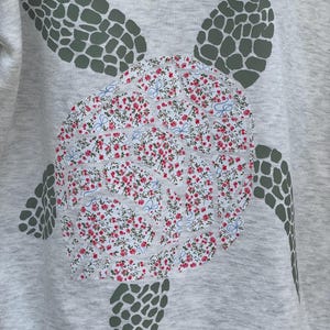 Puede incluir: Sudadera gris claro con un dise&ntilde;o de tortuga marina grande. El caparaz&oacute;n de la tortuga es un estampado floral con flores rojas y detalles azules. Las aletas y la cabeza de la tortuga son de color verde oscuro.