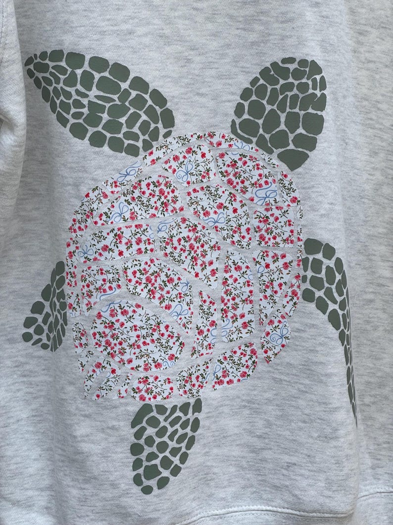 Puede incluir: Sudadera gris claro con un dise&ntilde;o de tortuga marina grande. El caparaz&oacute;n de la tortuga es un estampado floral con flores rojas y detalles azules. Las aletas y la cabeza de la tortuga son de color verde oscuro.