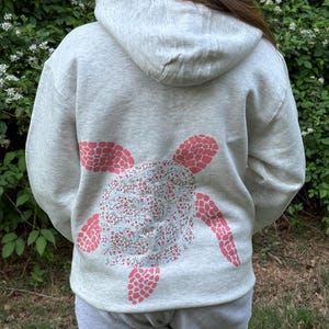Puede incluir: Sudadera con capucha gris claro con un dise&ntilde;o de tortuga en la espalda. El caparaz&oacute;n de la tortuga est&aacute; lleno de un estampado floral, y las aletas son de color rosa. La sudadera tiene capucha y bolsillo de canguro.