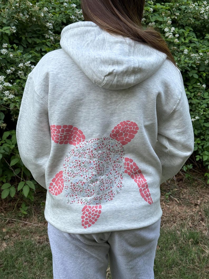 Puede incluir: Sudadera con capucha gris claro con un dise&ntilde;o de tortuga en la espalda. El caparaz&oacute;n de la tortuga est&aacute; lleno de un estampado floral, y las aletas son de color rosa. La sudadera tiene capucha y bolsillo de canguro.