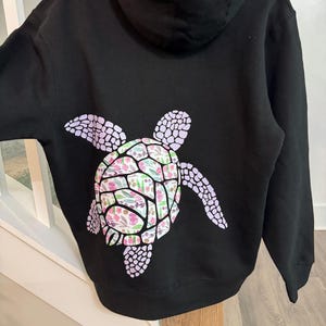 Puede incluir: Sudadera con capucha negra con un dise&ntilde;o de tortuga en la espalda. El caparaz&oacute;n de la tortuga tiene un estampado floral en rosa, verde y blanco. Las aletas de la tortuga son de color lila claro. Prenda casual y c&oacute;moda.