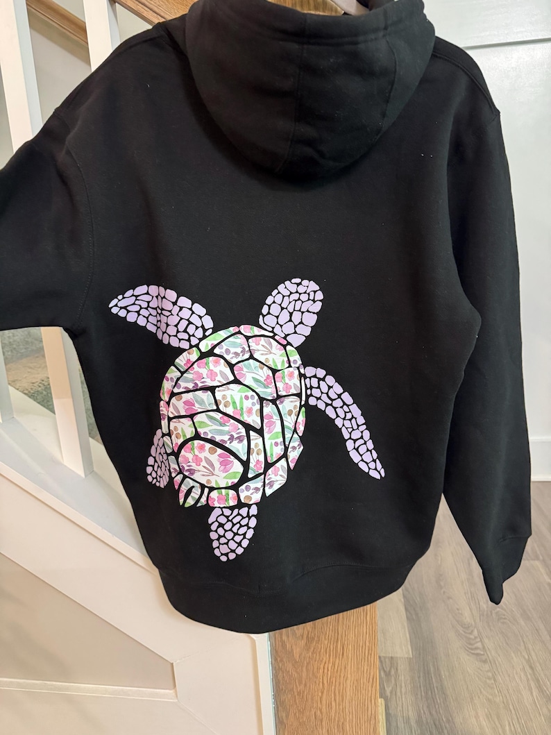 Puede incluir: Sudadera con capucha negra con un dise&ntilde;o de tortuga en la espalda. El caparaz&oacute;n de la tortuga tiene un estampado floral en rosa, verde y blanco. Las aletas de la tortuga son de color lila claro. Prenda casual y c&oacute;moda.