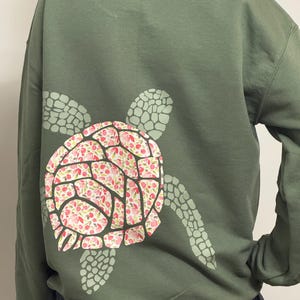 Puede incluir: Una sudadera verde oliva con un dise&ntilde;o de tortuga en la espalda. El caparaz&oacute;n de la tortuga est&aacute; decorado con motivos florales rosas y rojos, mientras que la cabeza y las aletas son de color verde claro.