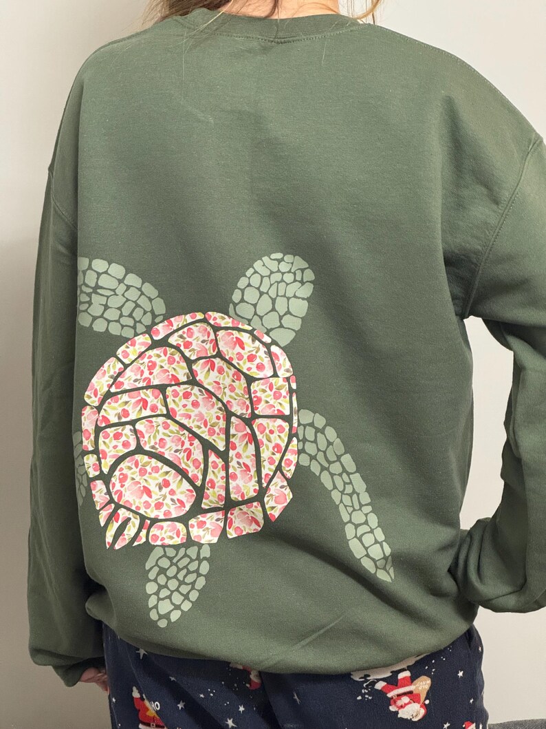 Puede incluir: Una sudadera verde oliva con un dise&ntilde;o de tortuga en la espalda. El caparaz&oacute;n de la tortuga est&aacute; decorado con motivos florales rosas y rojos, mientras que la cabeza y las aletas son de color verde claro.