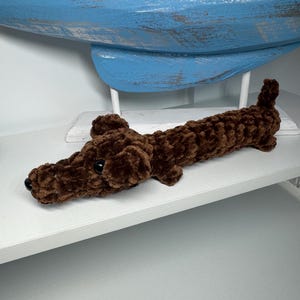 May include: A brown, plush dachshund dog toy with black eyes and nose. The toy is made of a soft, textured material and is lying on a white shelf. A blue wooden boat is in the background.