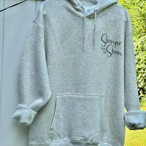 May include: A light gray heathered hoodie featuring a front kangaroo pocket, long sleeves with rolled cuffs, and a drawstring hood. The text "Stronger THAN THE Storm" is printed in black script and block lettering on the upper left chest area.