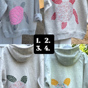 Puede incluir: Cuatro sudaderas con capucha grises con dise&ntilde;os de tortugas. Las tortugas tienen diferentes patrones y colores, incluyendo rosa, rojo, verde y amarillo. Las sudaderas se muestran sobre una superficie blanca.
