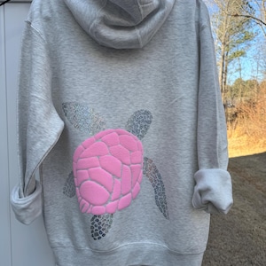 Puede incluir: Una sudadera con capucha gris claro con un dise&ntilde;o de tortuga rosa en la espalda. El caparaz&oacute;n de la tortuga es rosa y las aletas y la cabeza son plateadas con purpurina. La sudadera tiene capucha y mangas largas.