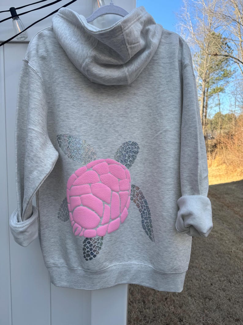 Puede incluir: Una sudadera con capucha gris claro con un dise&ntilde;o de tortuga rosa en la espalda. El caparaz&oacute;n de la tortuga es rosa y las aletas y la cabeza son plateadas con purpurina. La sudadera tiene capucha y mangas largas.