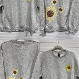 Puede incluir: Una sudadera gris con cuello redondo y mangas largas. La parte delantera presenta un bordado de girasol amarillo. La parte trasera de la sudadera tiene un dise&ntilde;o de girasol m&aacute;s grande con p&eacute;talos amarillos y un centro marr&oacute;n. La sudadera est&aacute; colgada de una percha.