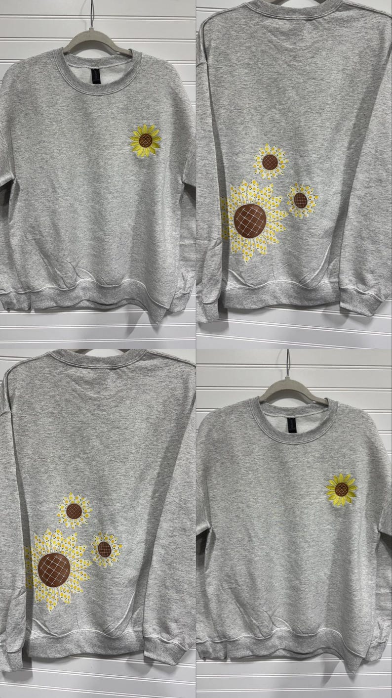 Puede incluir: Una sudadera gris con cuello redondo y mangas largas. La parte delantera presenta un bordado de girasol amarillo. La parte trasera de la sudadera tiene un dise&ntilde;o de girasol m&aacute;s grande con p&eacute;talos amarillos y un centro marr&oacute;n. La sudadera est&aacute; colgada de una percha.