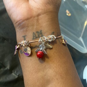 May include: A gold charm bracelet with a variety of charms, including a crescent moon, a red bead, and an angel wing charm. The bracelet is on a person's wrist.