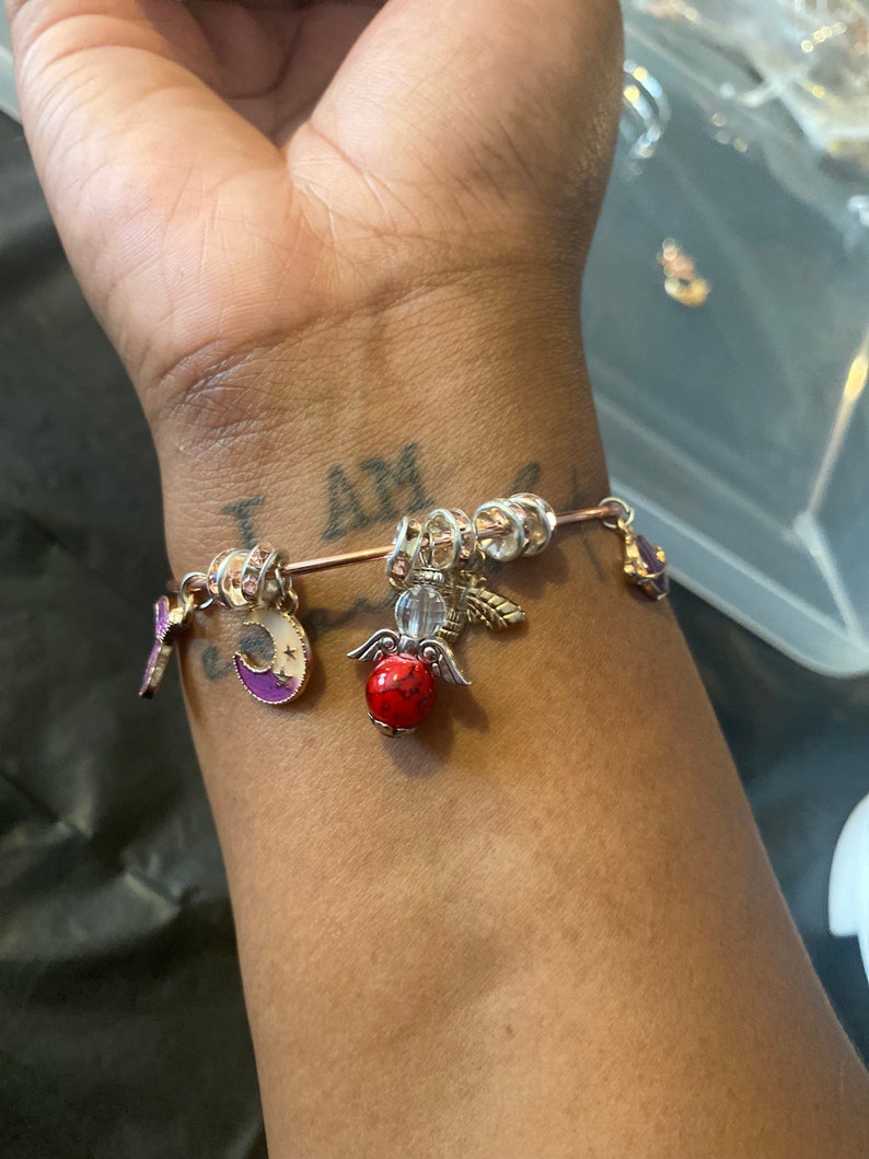 May include: A gold charm bracelet with a variety of charms, including a crescent moon, a red bead, and an angel wing charm. The bracelet is on a person's wrist.