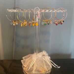 May include: A set of gold hoop earrings with various charms, including flowers, leaves, and beads. The earrings are displayed on a clear stand with a white organza bag.