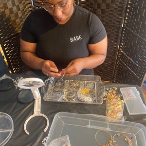 May include: A person is making jewelry with gold and silver hoops, beads, and charms. The jewelry is arranged in clear plastic containers on a black surface. The person is wearing a black shirt with the word "BABE" printed on it.