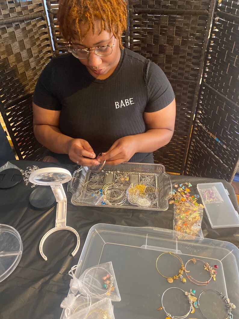 May include: A person is making jewelry with gold and silver hoops, beads, and charms. The jewelry is arranged in clear plastic containers on a black surface. The person is wearing a black shirt with the word "BABE" printed on it.