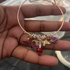 May include: A rose gold charm bracelet with a variety of charms, including a mermaid, a crescent moon, a bee, a crystal bead, and a purple planet. The bracelet is adorned with small, clear rhinestones.