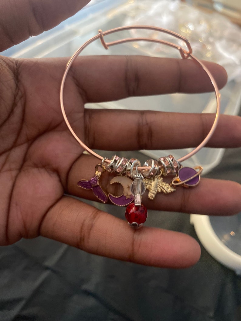 May include: A rose gold charm bracelet with a variety of charms, including a mermaid, a crescent moon, a bee, a crystal bead, and a purple planet. The bracelet is adorned with small, clear rhinestones.