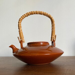 May include: A brown ceramic teapot with a woven wicker handle. The teapot has a rounded body and a wide, flat spout.