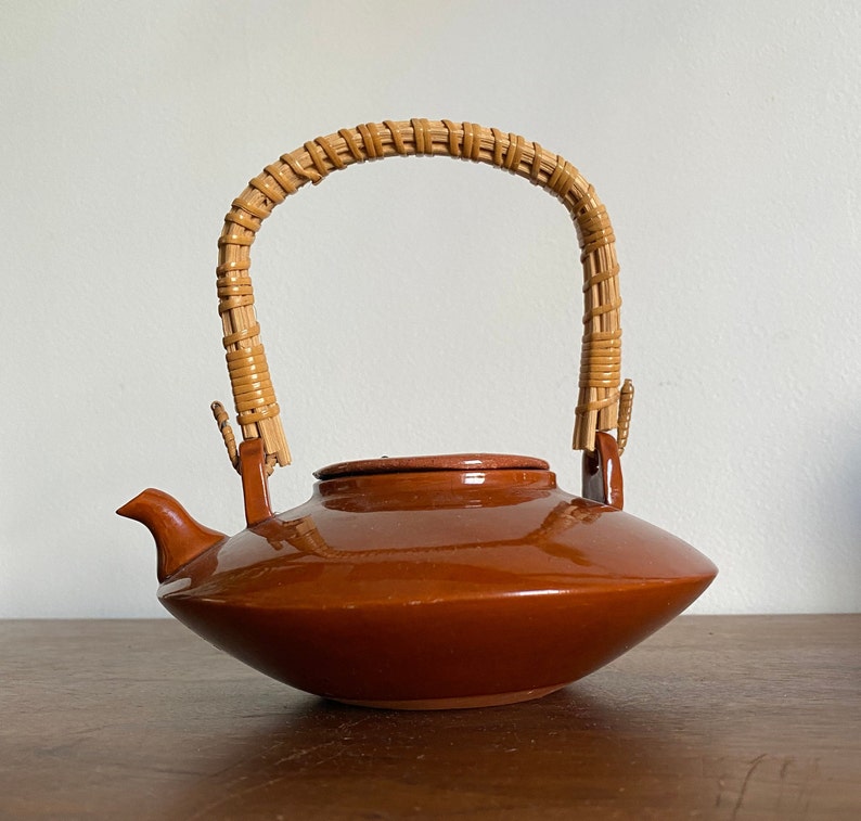 May include: A brown ceramic teapot with a woven wicker handle. The teapot has a rounded body and a wide, flat spout.