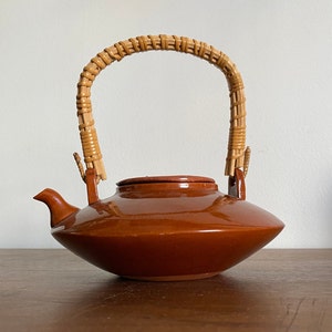 May include: A brown ceramic teapot with a woven wicker handle. The teapot has a rounded body and a wide, flat spout.