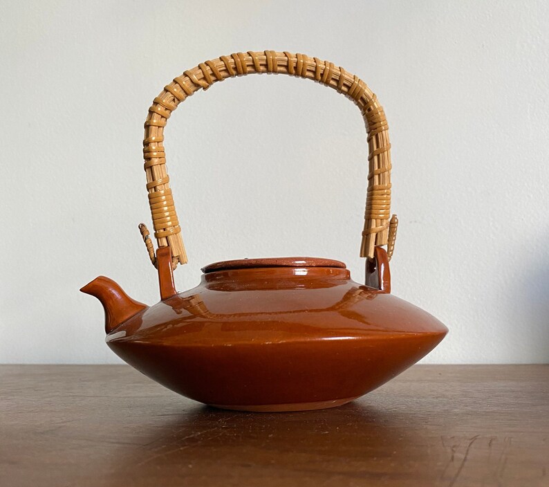 May include: A brown ceramic teapot with a woven wicker handle. The teapot has a rounded body and a wide, flat spout.