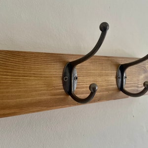 May include: A wooden wall-mounted coat rack with two metal hooks. The rack is stained a dark brown color and the hooks are a dark gray metal.
