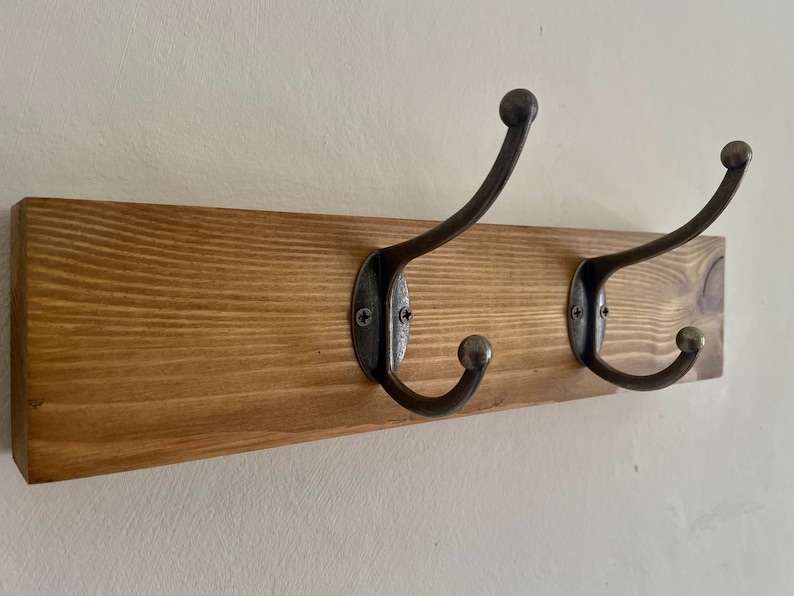 May include: A wooden wall-mounted coat rack with two metal hooks. The rack is stained a dark brown color and the hooks are a dark gray metal.
