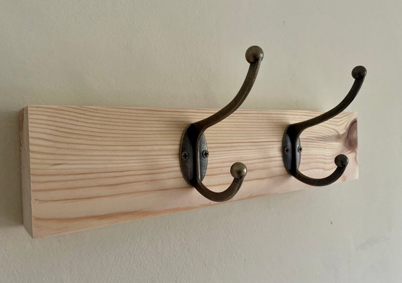 May include: A wooden coat rack with two antique brass hooks. The rack is made of natural wood with a light brown finish.