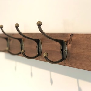 May include: A wooden wall-mounted coat rack with three antique brass hooks. The rack is stained a dark brown colour.
