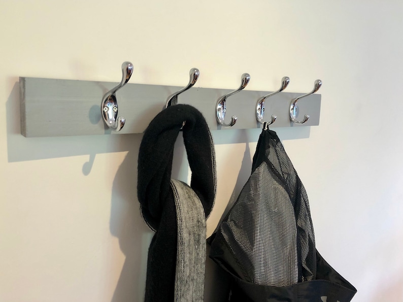K&ouml;nnte beinhalten: Ein grauer, an der Wand montierter Kleiderst&auml;nder mit f&uuml;nf verchromten Haken. Ein schwarz-grauer Schal und eine schwarze Netztasche h&auml;ngen an den Haken.