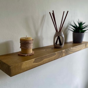 May include: A wooden shelf with a natural finish, mounted on a white wall. The shelf is decorated with a small candle, a diffuser with reeds, and a potted succulent plant.