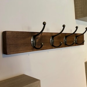 May include: A wooden wall-mounted coat rack with four metal hooks. The rack is stained a dark brown color and the hooks are a dark bronze color.