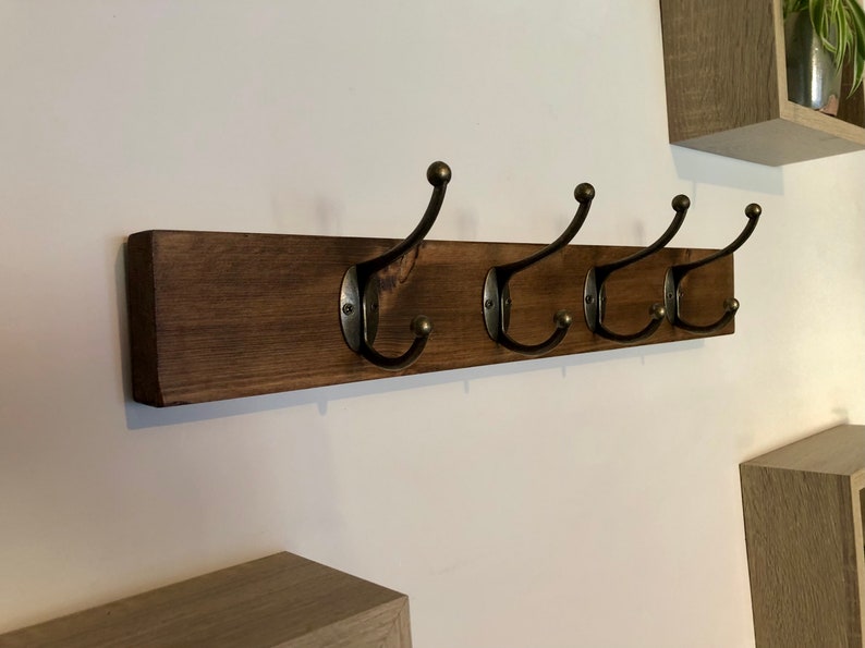 May include: A wooden wall-mounted coat rack with four metal hooks. The rack is stained a dark brown color and the hooks are a dark bronze color.