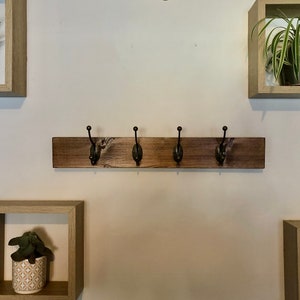May include: A wooden coat rack with four hooks, mounted on a white wall. The rack is stained a dark brown color. There are three square shelves with plants on the wall.