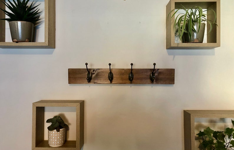 May include: A wooden coat rack with four hooks, mounted on a white wall. The rack is stained a dark brown color. There are three square shelves with plants on the wall.