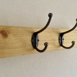 May include: A wooden wall-mounted coat rack with two black metal hooks. The rack is made of natural wood with a light brown finish.