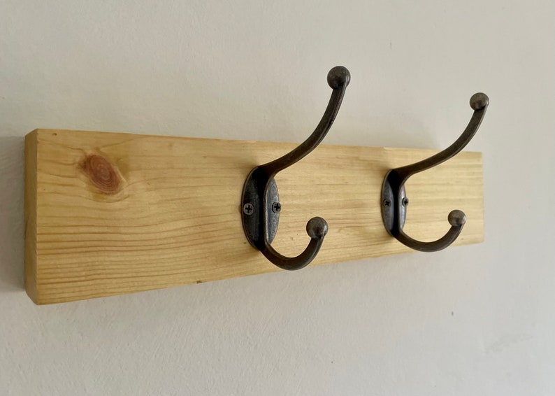 May include: A wooden wall-mounted coat rack with two black metal hooks. The rack is made of natural wood with a light brown finish.