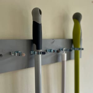 May include: A gray wooden wall-mounted organizer with metal clips holds three cleaning tools. The tools include a gray mop handle, a white mop handle, and a green mop handle.