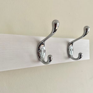 K&ouml;nnte beinhalten: Ein wei&szlig;er, an der Wand montierter Kleiderst&auml;nder mit zwei verchromten Haken.