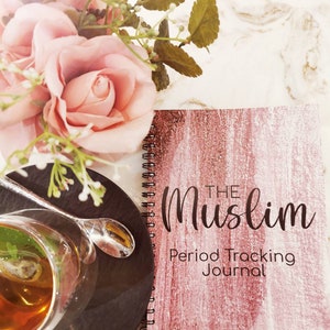 May include: A pink and white journal with the text "The Muslim Period Tracking Journal" on the cover. The journal is lying on a white surface with a cup of tea and a bouquet of pink roses in the background.