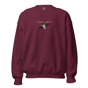 May include: A maroon sweatshirt with the text "I don't spit, I" above a graphic of a bird in flight. The bird has a white body, brown wings, and a yellow head. The sweatshirt has a crew neck and long sleeves.