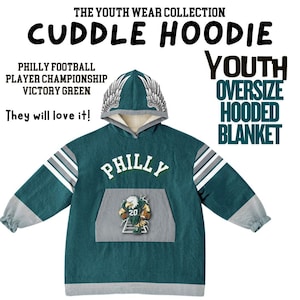 May include: A teal oversized hooded blanket with the word "PHILLY" in white lettering. The blanket features a hood with wing accents and a kangaroo pocket with a football player graphic. The text "THE YOUTH WEAR COLLECTION" is at the top.