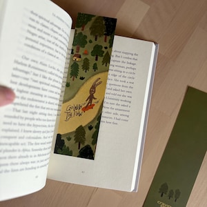 May include: A rectangular bookmark featuring a forest illustration with a brown rabbit on a skateboard and the text "Go With The Flow" in black ink. A second solid olive green bookmark with a small tree graphic and the text "cravin' space" is visible.