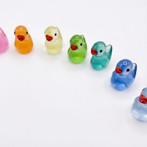 May include: A collection of small, translucent glass duck figurines arranged in a curved line. Each duck features a smooth, glossy finish, a distinct vibrant color, a small black eye, and a bright red beak, showcasing a spectrum of hues.